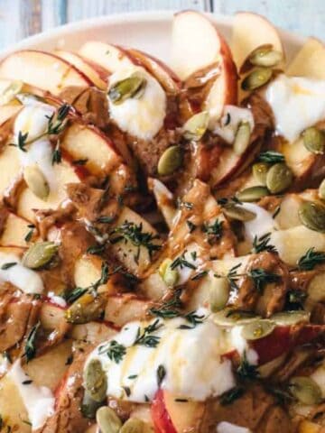 A plate of sliced apples topped with dollops of yogurt, drizzles of almond butter, sprinkled pumpkin seeds, and fresh thyme leaves, displayed on a rustic wooden surface.