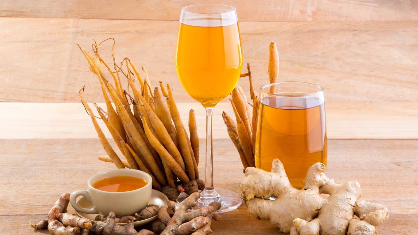 A glass of orange juice, a cup of orange liquid, and a glass of amber liquid are arranged on a wooden surface. Fresh ginger roots and long brown roots are placed around the beverages.