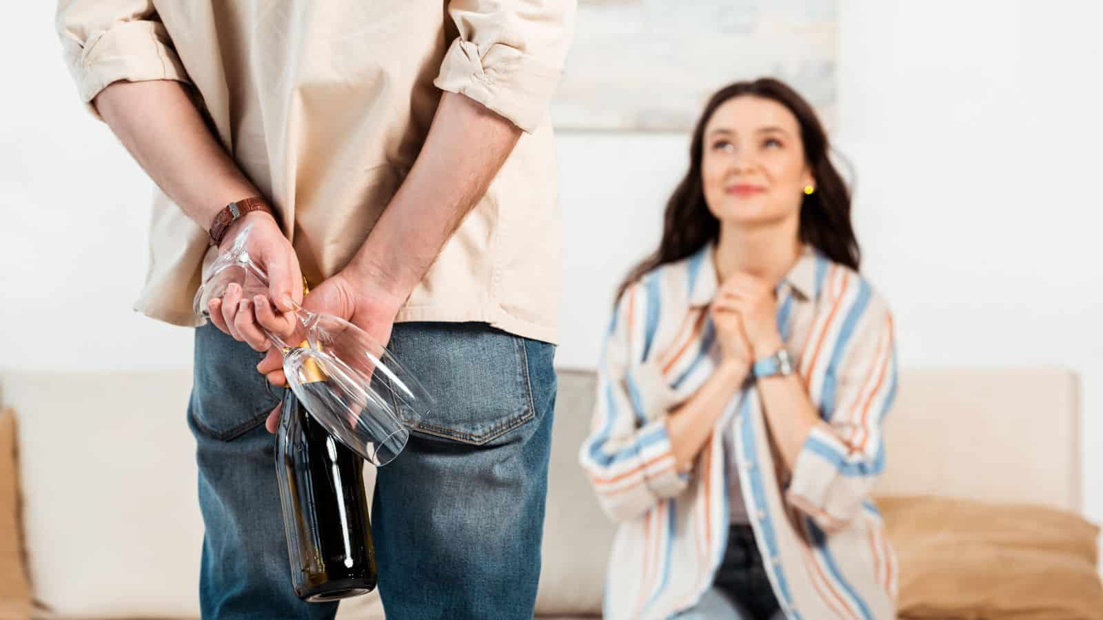 A man stands with his back to the viewer, holding a bottle of wine and two glasses behind him. A woman sits on a sofa, smiling and clasping her hands together. Both are in a warmly lit room.