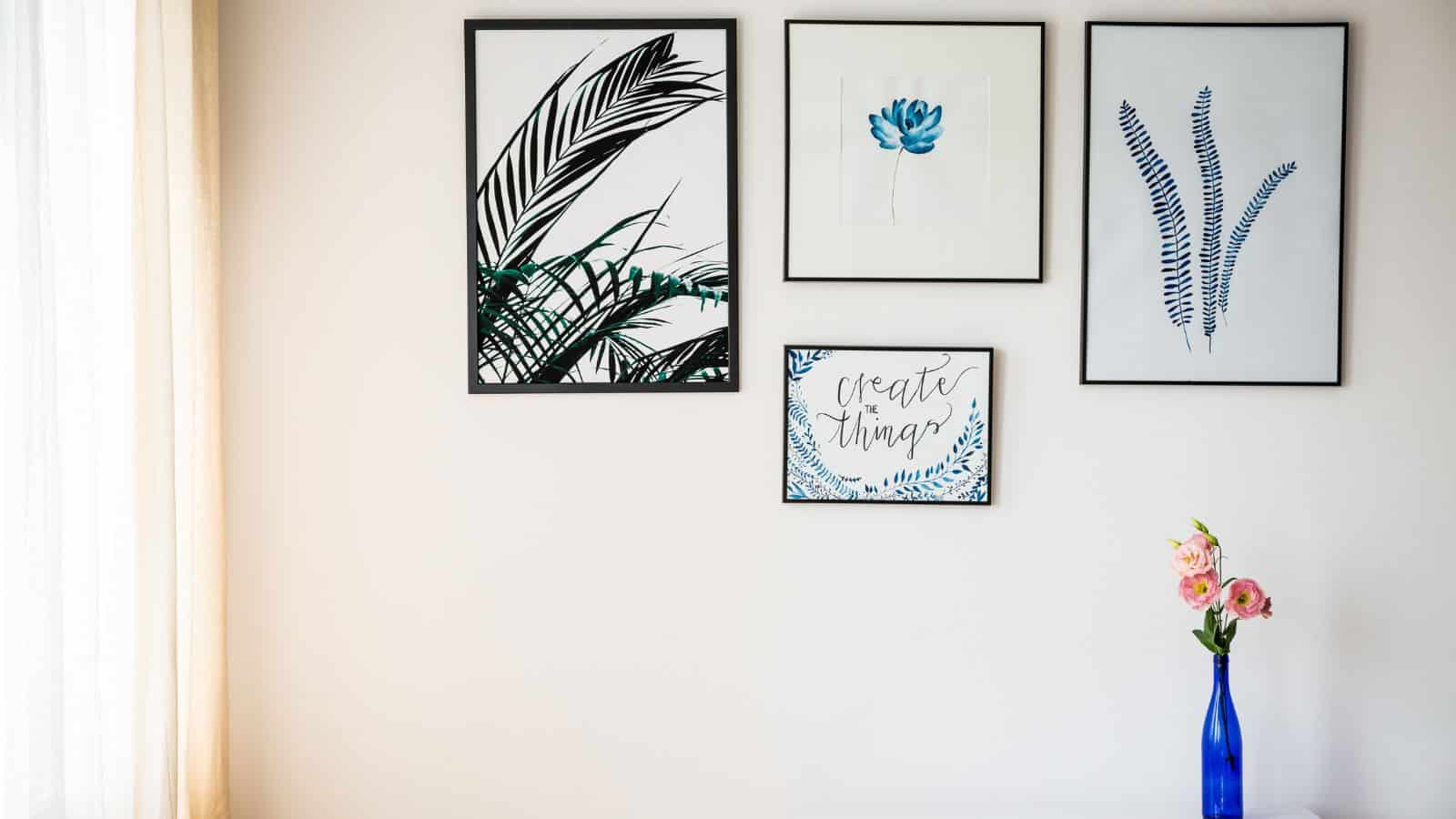 A white wall with four framed artworks. Three are botanical prints: two with leaves and one with a blue flower. The fourth frame has the text "create things" with floral accents. A small vase with pink flowers sits on a table below.