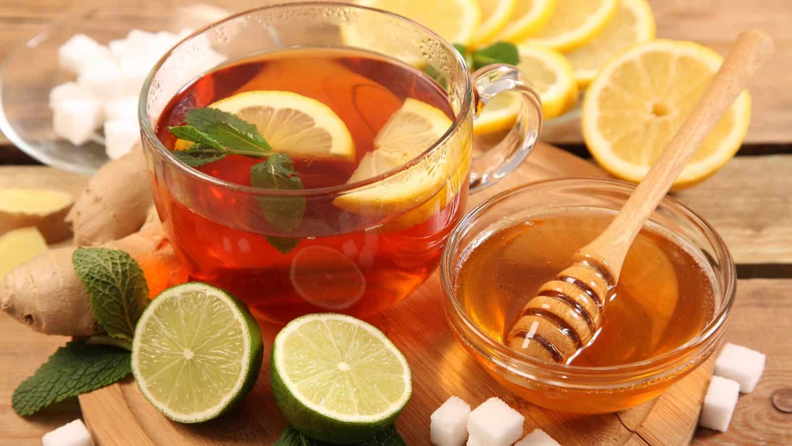 A cup of tea with lemon and mint sits on a wooden board next to a bowl of honey with a dipper. Fresh lime halves, lemon slices, ginger, mint leaves, and sugar cubes are arranged around the tea and honey.