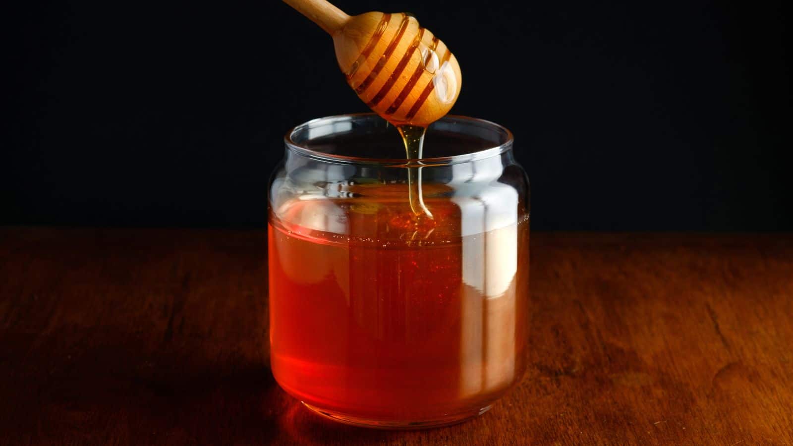 A wooden honey dipper drips honey into a clear glass jar filled with honey, set on a wooden surface with a dark background.