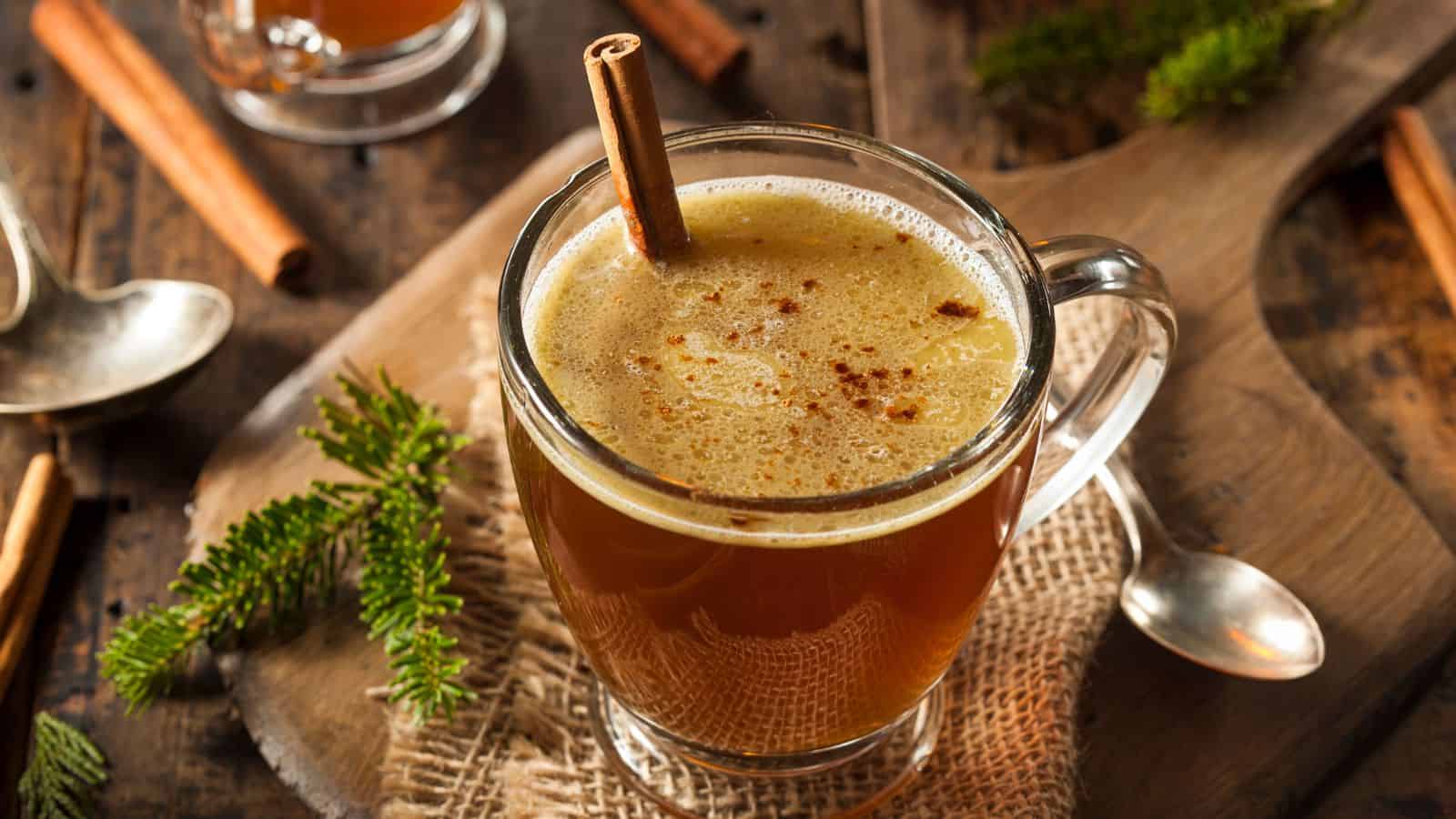 A glass mug filled with spiced hot apple cider garnished with a cinnamon stick. The mug sits on a wooden surface with small pine branches, cinnamon sticks, and a metal spoon nearby.