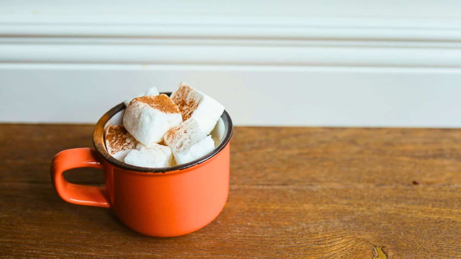 An orange mug filled with hot chocolate topped with marshmallows and a sprinkle of cocoa powder sits on a wooden surface. A white wall serves as the background.