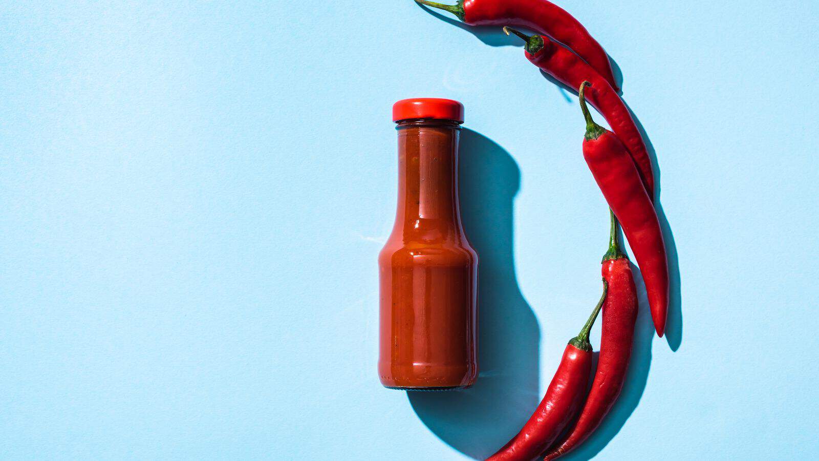 A red hot sauce bottle is placed against a light blue background. Four red chili peppers are arranged in a curved line on the right side of the bottle. The scene is brightly lit, casting soft shadows.