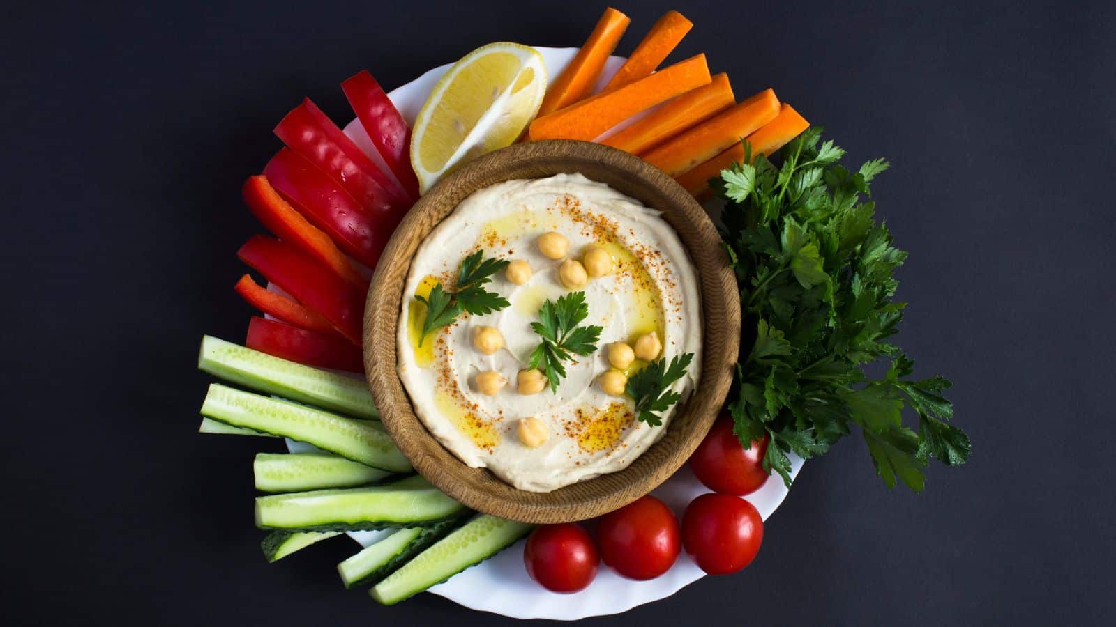 A bowl of hummus topped with chickpeas and herbs is surrounded by red bell pepper strips, carrot sticks, cucumber sticks, cherry tomatoes, fresh parsley, and a lemon wedge, arranged on a white plate against a dark background.