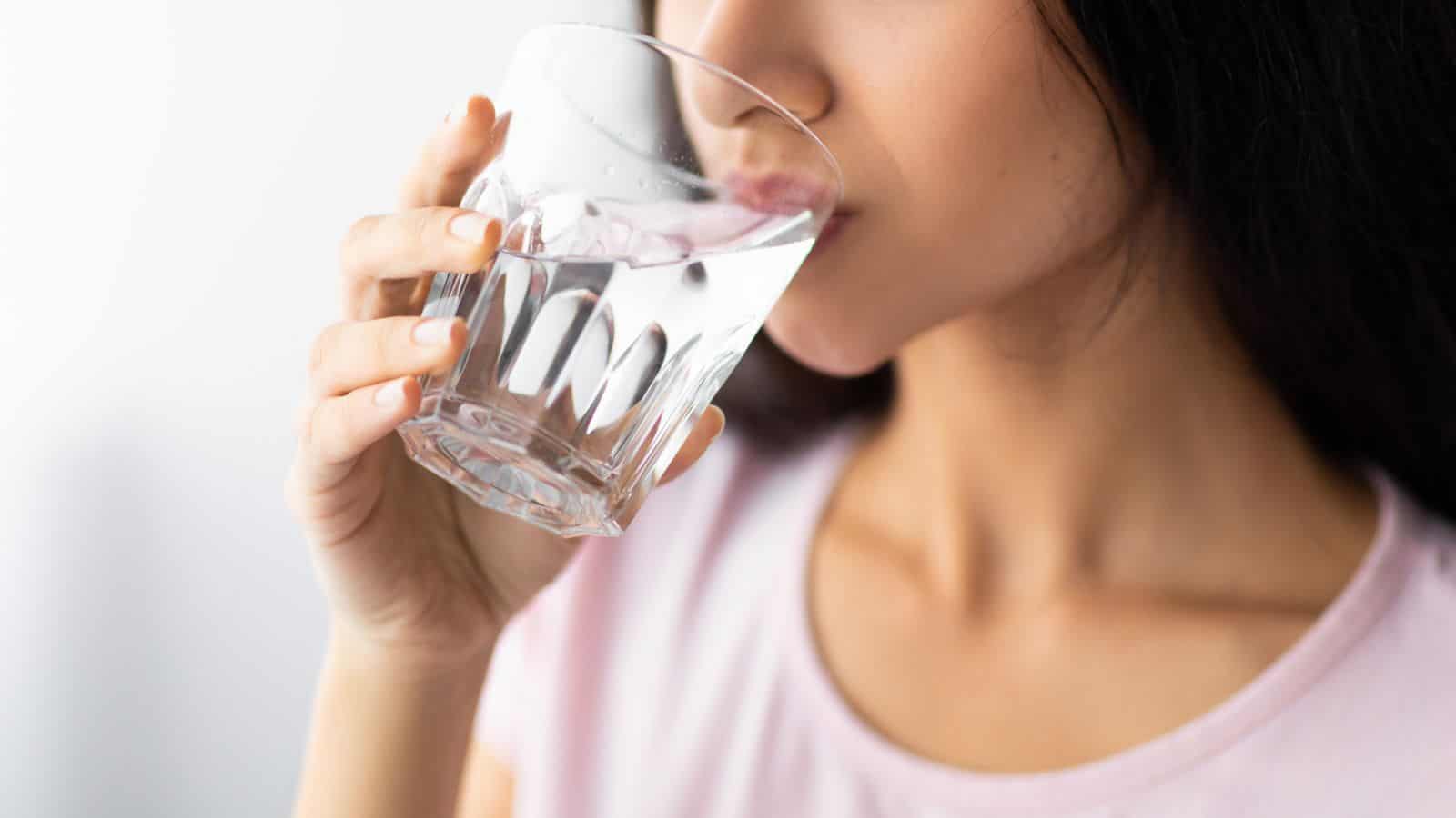 A woman in a pink shirt drinks water from a clear glass. Her face is partially visible, and she holds the glass with her right hand. The background is a light, neutral color.