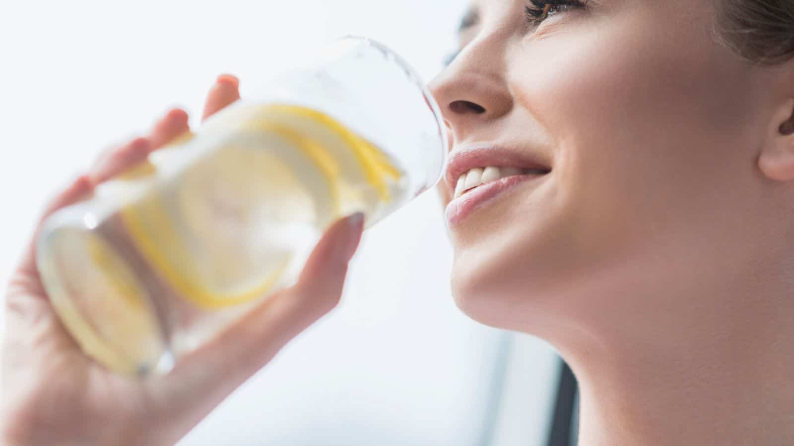 Close-up of a person drinking a glass of water with lemon slices. The background is softly blurred, and the lighting is bright, highlighting the freshness of the drink. The person appears to be smiling, enjoying the beverage.