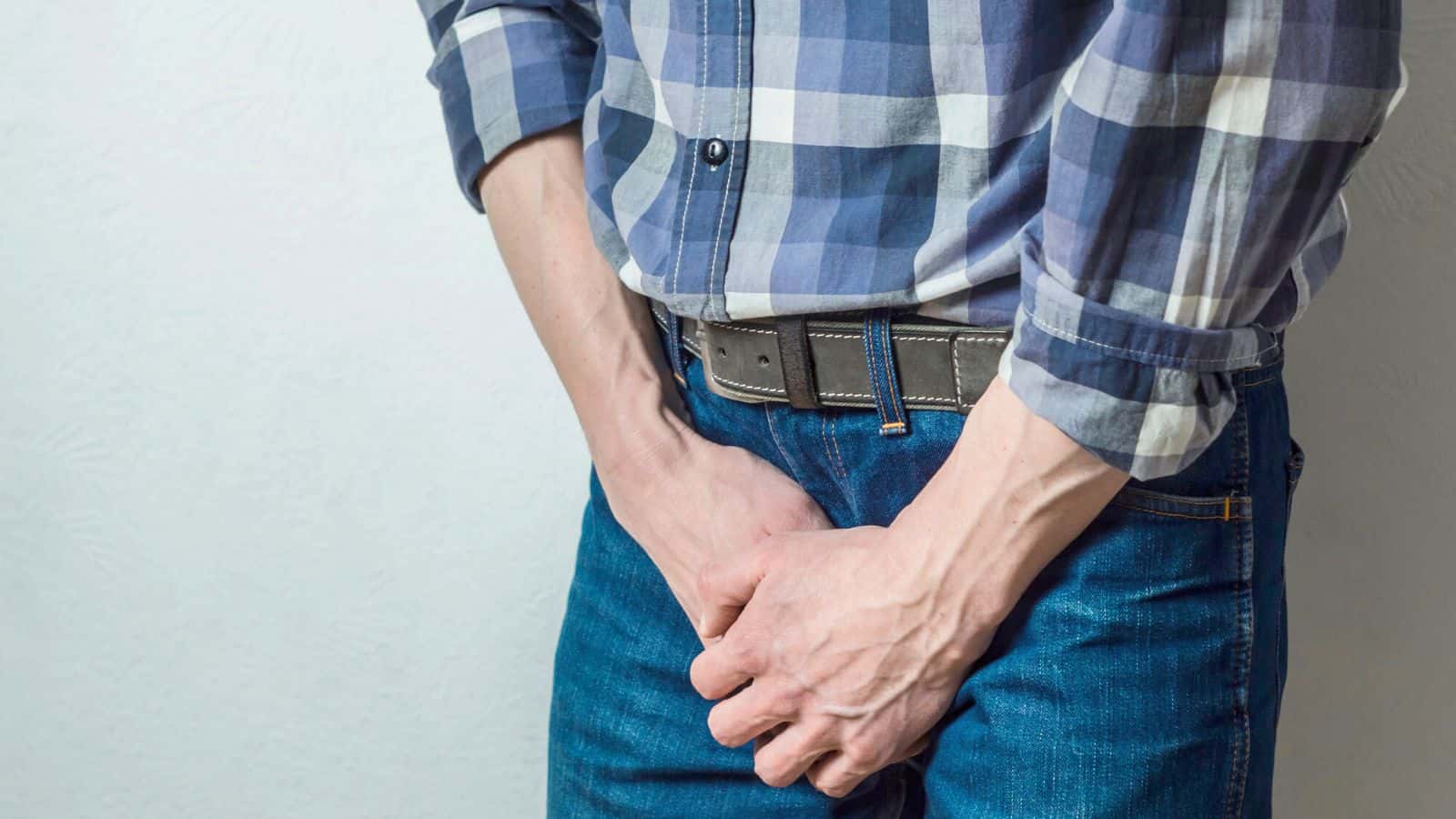 A person in a checkered shirt and jeans is standing against a white background, clutching their hands in front of the lower torso.