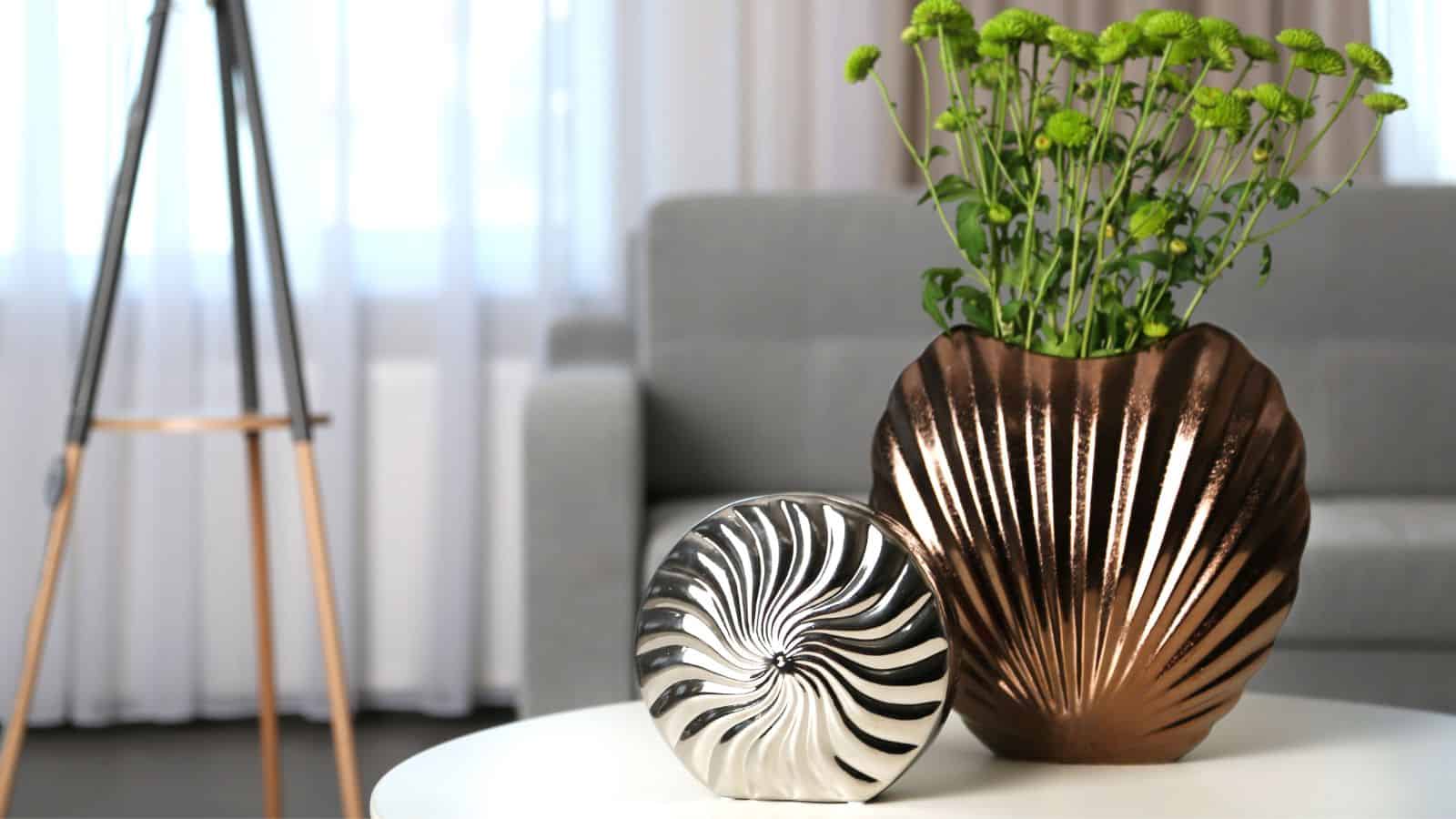 A modern living room features a metallic vase with green flowers and a silver decorative object on a white table. A grey sofa and a tripod-style lamp are in the background, with sheer curtains allowing natural light to filter in.