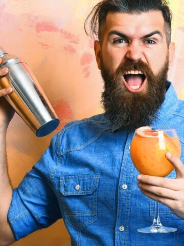 A bearded man in a blue shirt enthusiastically shakes a cocktail shaker while holding a glass with an orange drink. The background is colorful with pink and orange hues, and two other drinks are placed on the table in front of him.