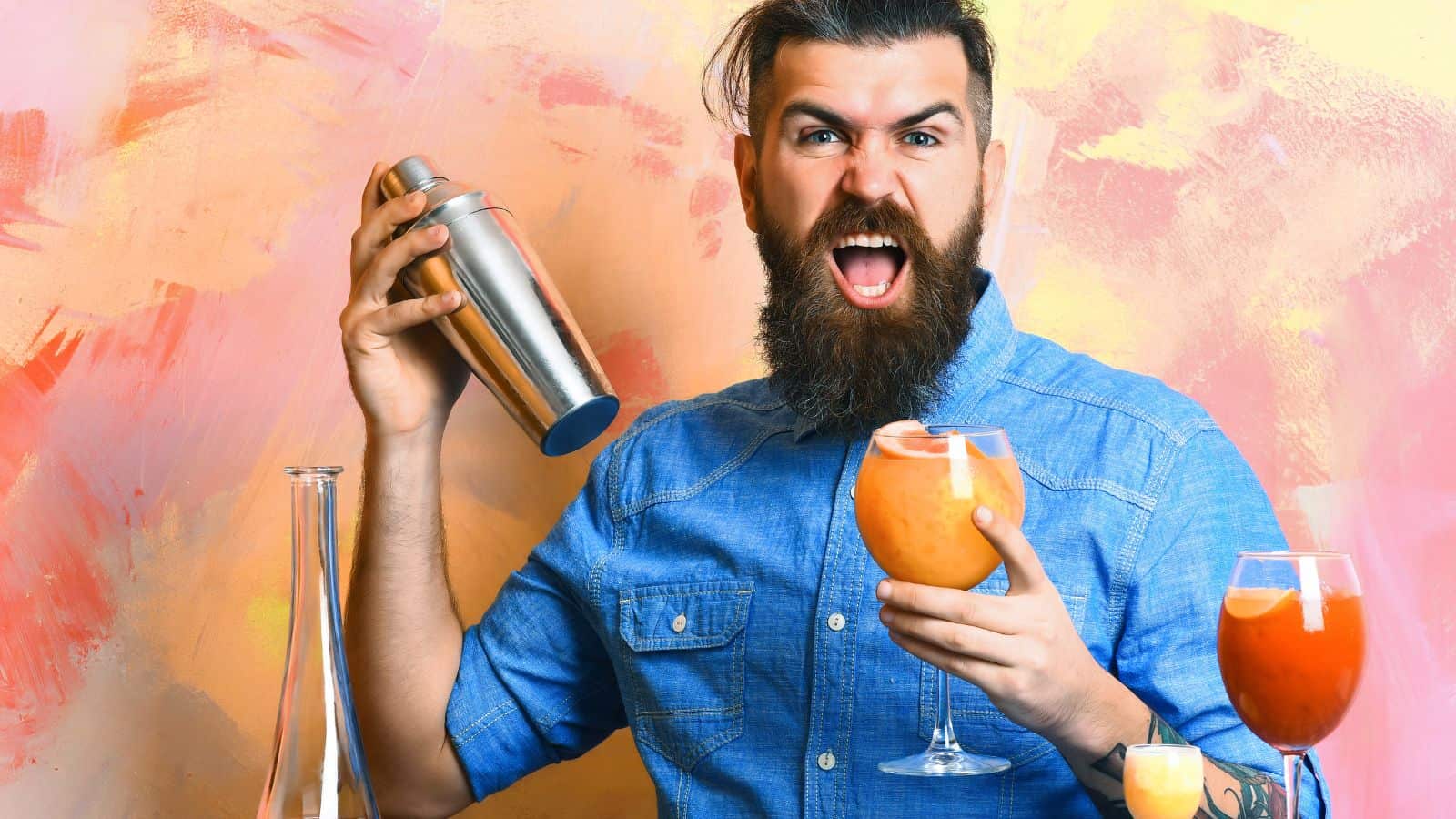 Man with a beard and tattoos shakes a cocktail shaker while holding a cocktail glass. He is wearing a blue denim shirt. There are two other filled cocktail glasses on a table. The background is painted in shades of pink and orange.