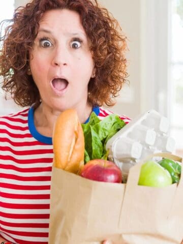 A person with curly hair and a surprised expression holds a brown paper bag filled with groceries, including a baguette, leafy greens, eggs, a red apple, and a green apple. They are wearing a red and white striped shirt. Bright windows are in the background.
