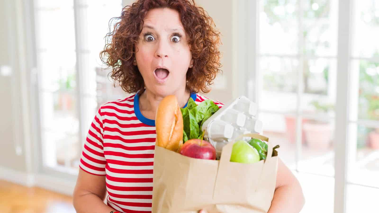 A person with curly hair and a surprised expression holds a brown paper bag filled with groceries, including a baguette, leafy greens, eggs, a red apple, and a green apple. They are wearing a red and white striped shirt. Bright windows are in the background.