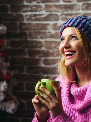 A person wearing a pink sweater and a colorful knit hat is holding a green mug and smiling. They are sitting in front of a decorated Christmas tree with red baubles and a gold bow, against a brick wall background.