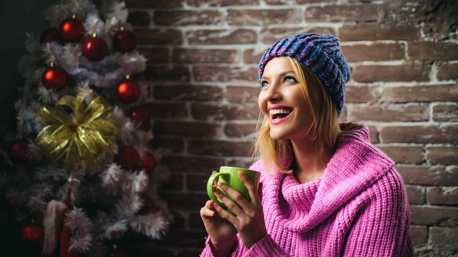 A person wearing a pink sweater and a colorful knit hat is holding a green mug and smiling. They are sitting in front of a decorated Christmas tree with red baubles and a gold bow, against a brick wall background.