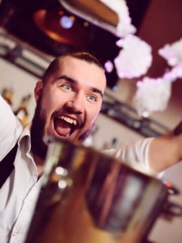 A bartender with tattoos energetically pours a drink, creating a splash above a cocktail shaker. He's bearded and sports a white shirt with suspenders. Amidst the warmly lit bar and shelves of bottles, he's breaking mocktail rules you need to challenge for sensational sips.