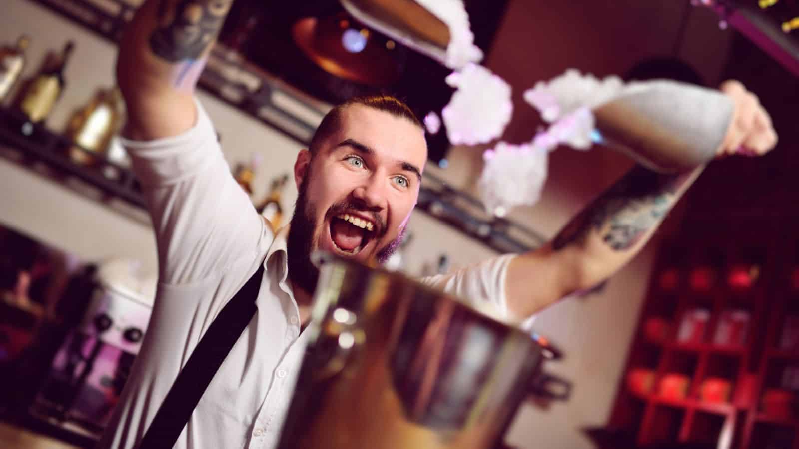 A bartender with tattoos energetically pours a drink, creating a splash above a cocktail shaker. He's bearded and sports a white shirt with suspenders. Amidst the warmly lit bar and shelves of bottles, he's breaking mocktail rules you need to challenge for sensational sips.