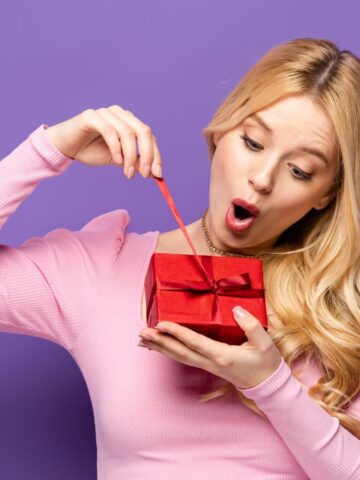 A woman in a pink sweater looks surprised while opening a small red gift box against a purple background.