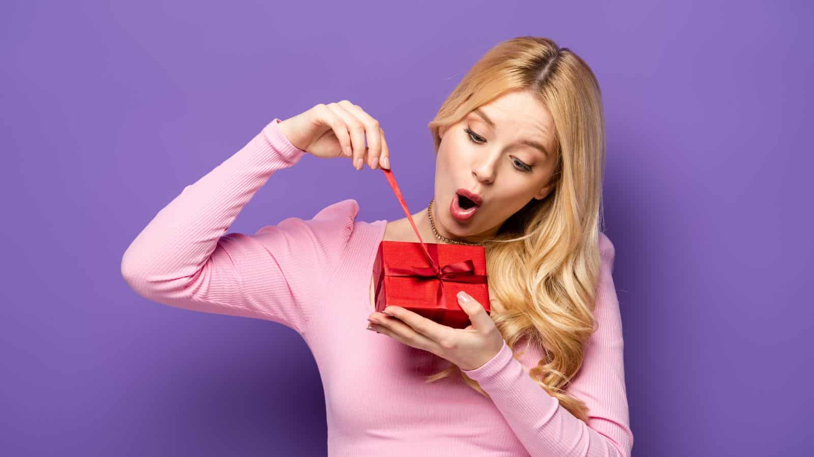 A woman in a pink sweater looks surprised while opening a small red gift box against a purple background.
