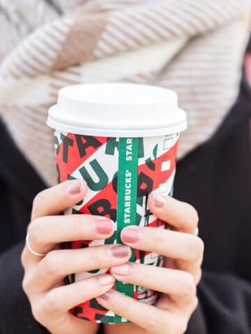 A person wearing a scarf and a dark jacket holds a festive Starbucks cup, brimming with hot, steamy goodness, in both hands. The cup features a white lid and vibrant red and green patterns with "Starbucks" visible. A pink backpack adds a splash of color to the cozy scene.
