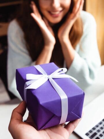 A person is holding a purple gift box with a white ribbon, extending it towards a woman who appears surprised. She is seated at a table with an open laptop and a cup nearby.