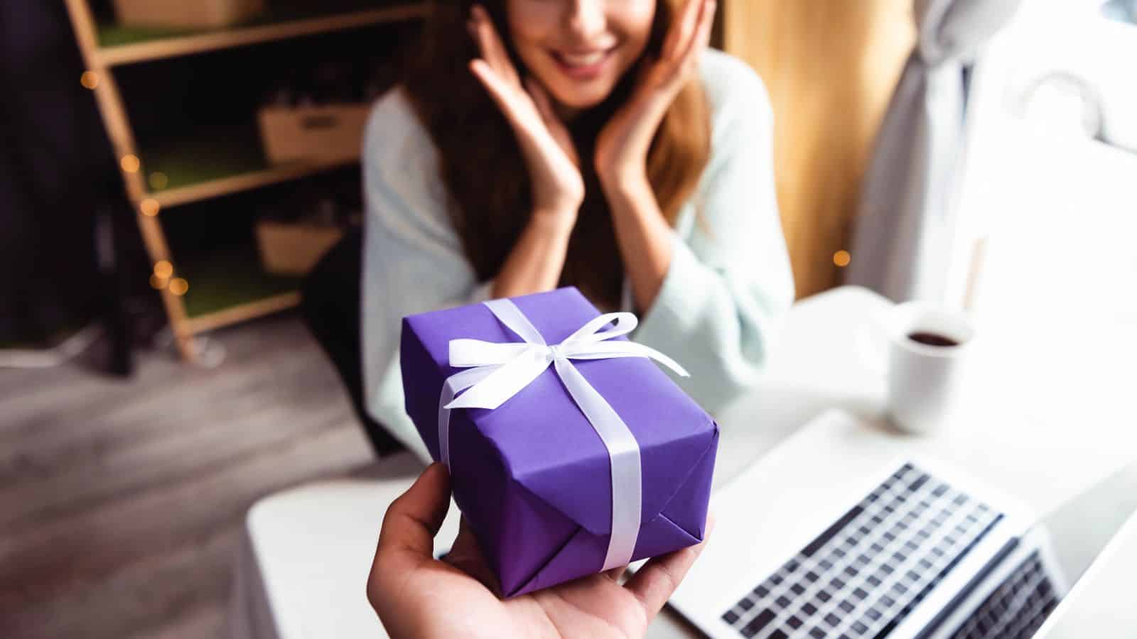A person is holding a purple gift box with a white ribbon, extending it towards a woman who appears surprised. She is seated at a table with an open laptop and a cup nearby.