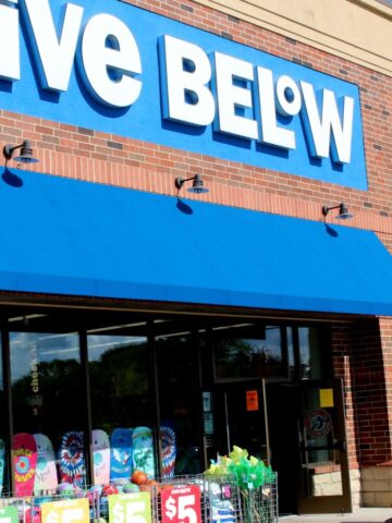 A store with a sign reading "Five Below" is visible. Blue awnings cover the entrance. Colorful products are displayed outside, with signs indicating a $5 price point. The building exterior is red brick, and the sky is partly cloudy.