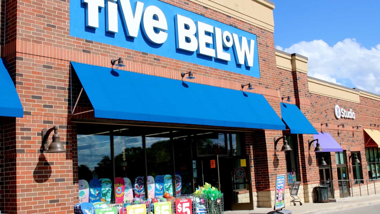 A store with a sign reading "Five Below" is visible. Blue awnings cover the entrance. Colorful products are displayed outside, with signs indicating a $5 price point. The building exterior is red brick, and the sky is partly cloudy.