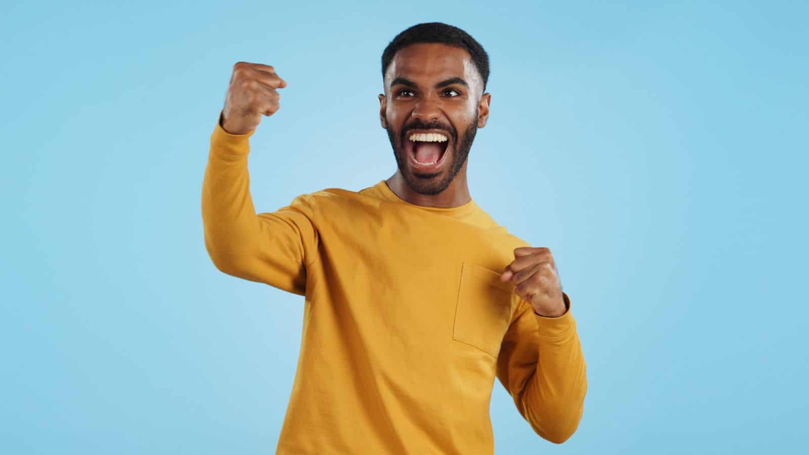 A person wearing a yellow long-sleeve shirt is celebrating with both hands raised and a wide smile against a light blue background.