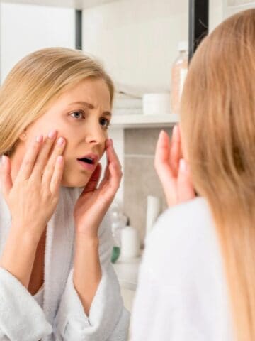 A woman with long blonde hair is standing in front of a bathroom mirror, wearing a white robe. She appears to be examining her face closely, touching her cheeks with a concerned expression. Toiletries are visible in the background.