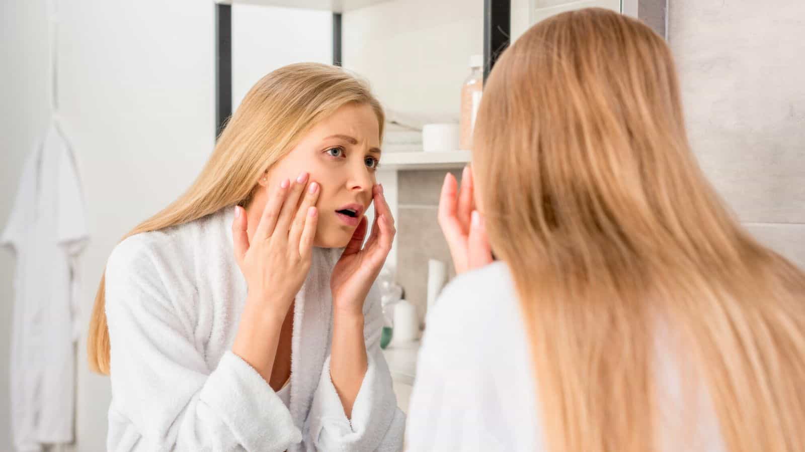 A woman with long blonde hair is standing in front of a bathroom mirror, wearing a white robe. She appears to be examining her face closely, touching her cheeks with a concerned expression. Toiletries are visible in the background.