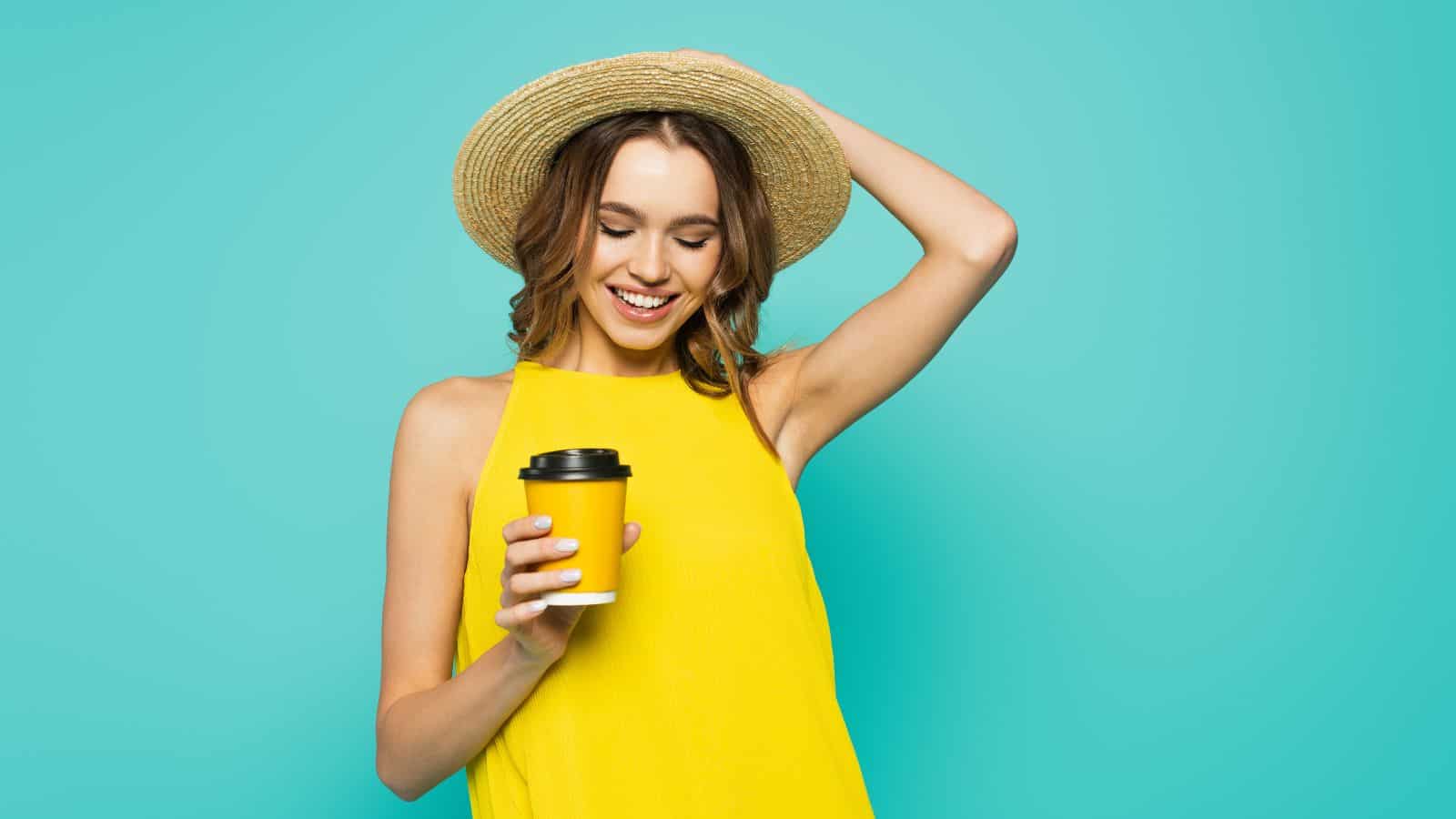 A person wearing a straw hat and a yellow sleeveless top smiles while holding a takeaway coffee cup against a turquoise background.