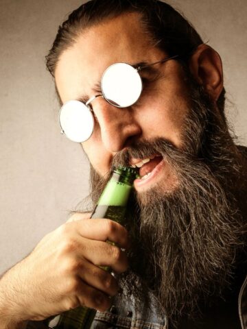 A bearded man with long hair wears reflective round sunglasses and a denim vest. He is attempting to open a green beer bottle with his teeth against a neutral background.