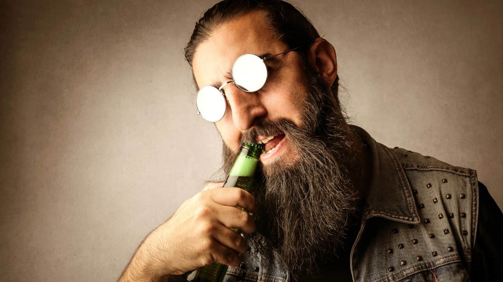 A bearded man with long hair wears reflective round sunglasses and a denim vest. He is attempting to open a green beer bottle with his teeth against a neutral background.