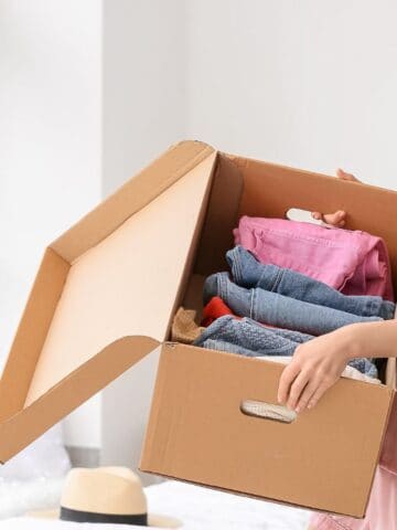 A woman in pink pajamas holds an open cardboard box filled with folded clothes. She stands in a bright room near a window. The bed behind her has a hat and pillows. The room has light-colored walls and decor.