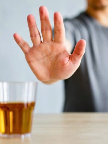 A person in a gray shirt extends their hand in a stop gesture towards a glass filled with a brown liquid and a bottle on a table. The image suggests refusal or rejection of an alcoholic beverage.