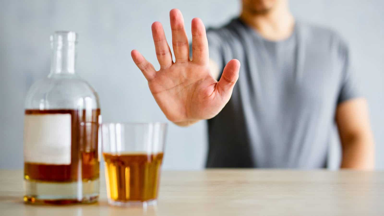 A person in a gray shirt extends their hand in a stop gesture towards a glass filled with a brown liquid and a bottle on a table. The image suggests refusal or rejection of an alcoholic beverage.