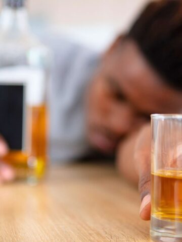 A person is resting their head on a wooden table, holding a glass of amber liquid, possibly whiskey, in the foreground. A bottle of similar liquid is nearby. The scene suggests relaxation or tiredness.