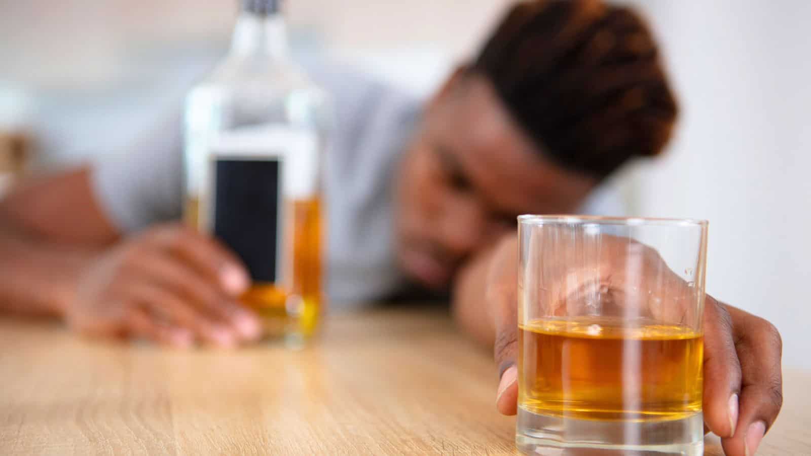 A person is resting their head on a wooden table, holding a glass of amber liquid, possibly whiskey, in the foreground. A bottle of similar liquid is nearby. The scene suggests relaxation or tiredness.