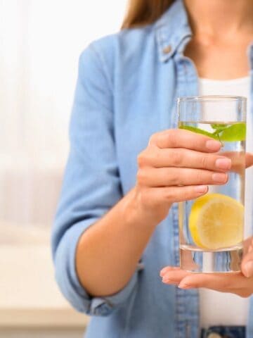 A person in a denim shirt holds a glass of water infused with lemon slices and a sprig of mint. The background is softly blurred, showing a light-colored room with furniture.