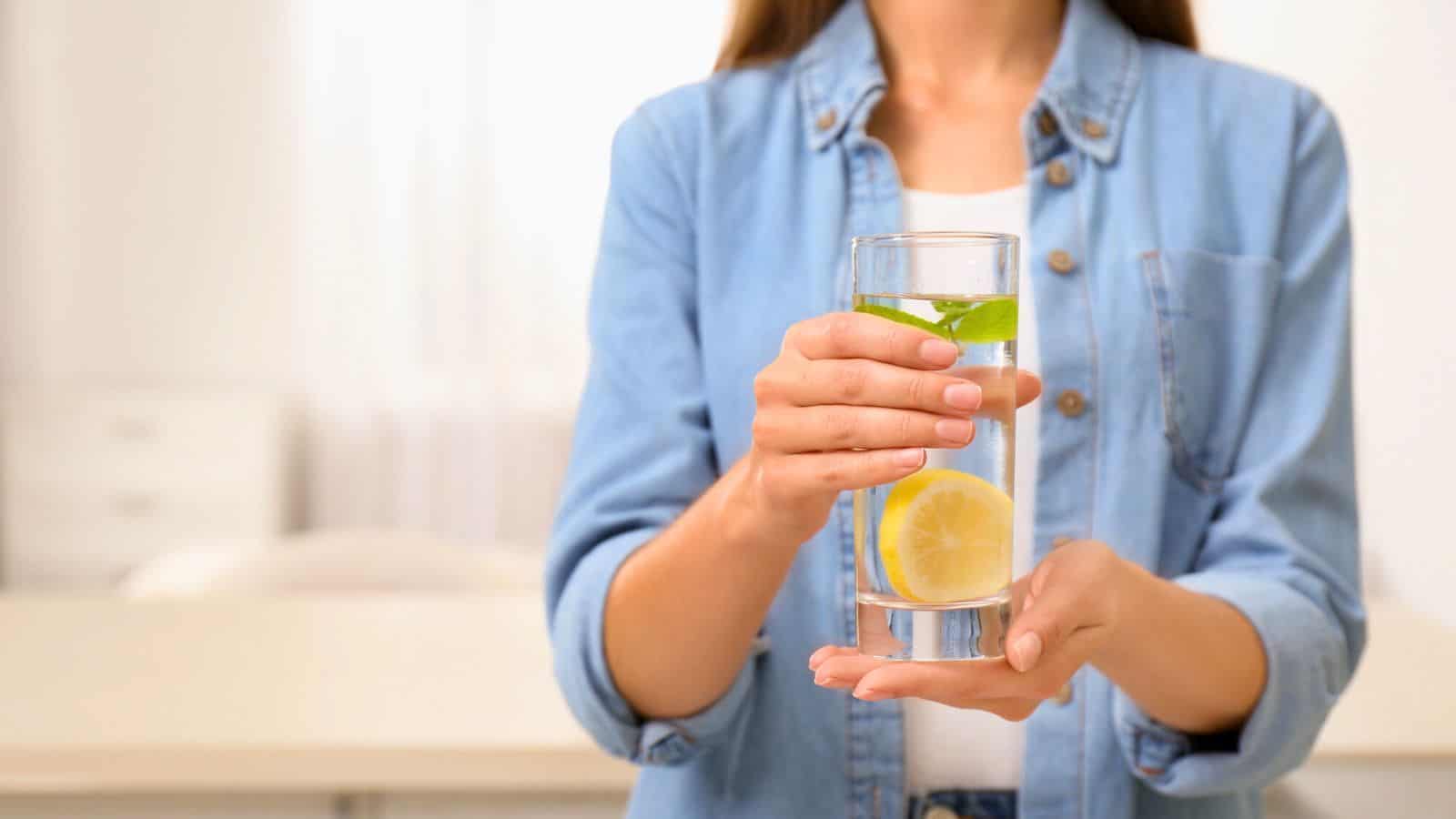 A person in a denim shirt holds a glass of water infused with lemon slices and a sprig of mint. The background is softly blurred, showing a light-colored room with furniture.