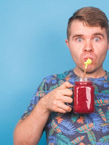 A person with wide eyes drinks a red beverage from a jar using a yellow straw. They are wearing a colorful, patterned shirt and are standing against a blue background.