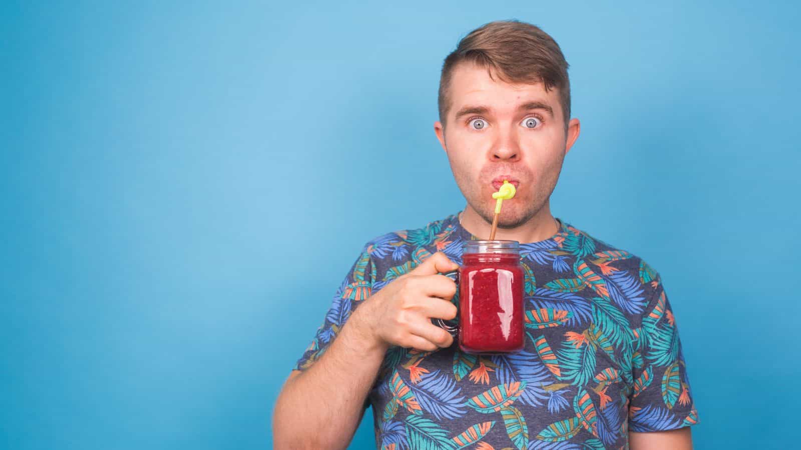 A person with wide eyes drinks a red beverage from a jar using a yellow straw. They are wearing a colorful, patterned shirt and are standing against a blue background.
