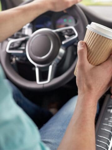 Two people in a car holding takeaway coffee cups. The driver is holding the steering wheel with one hand. The car's dashboard and center console are visible, showing various controls. The cups are in brown, ribbed, disposable holders.