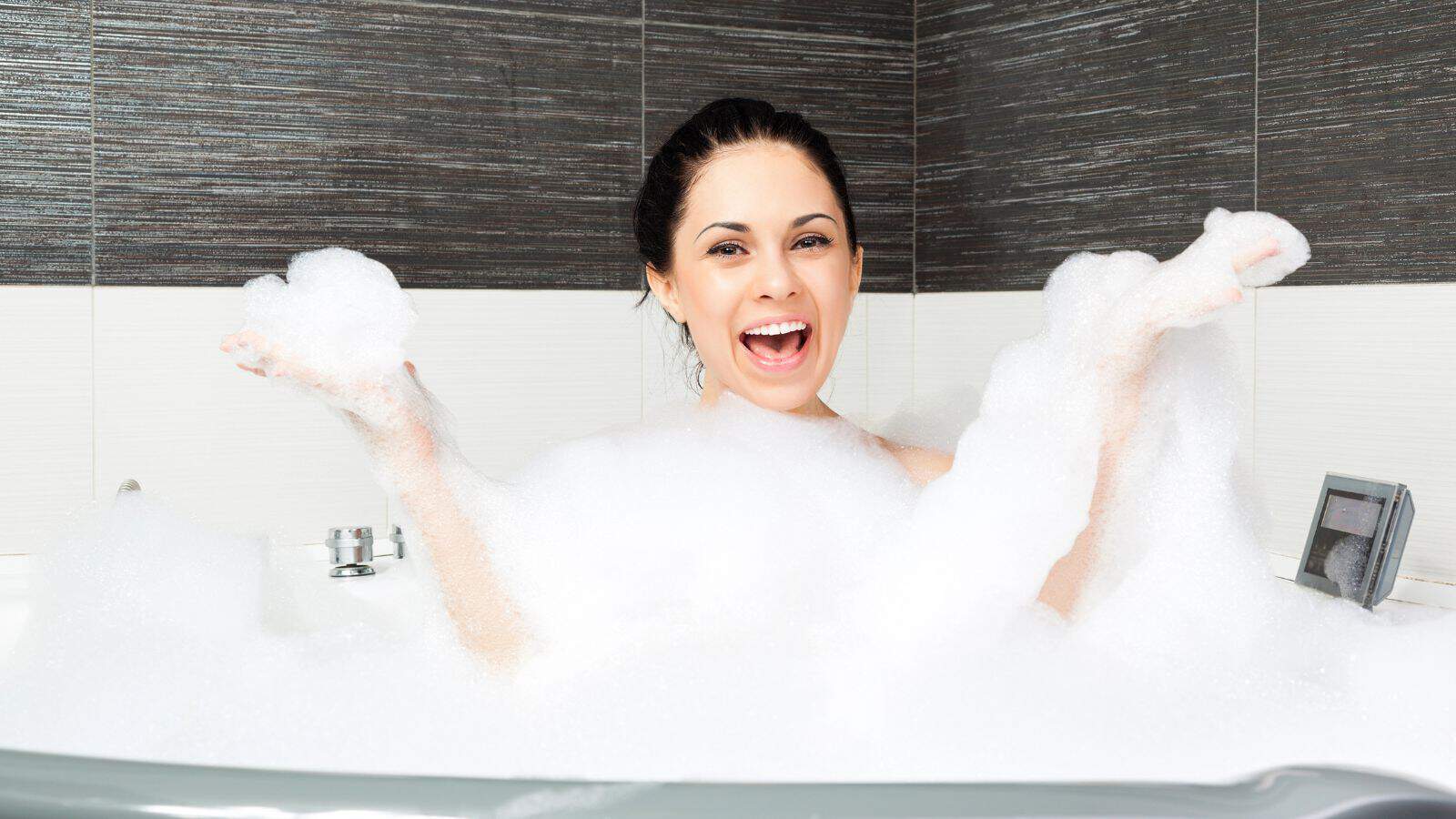 A person enjoys a bubble bath in a modern tiled bathroom, surrounded by lots of foam. Smiling broadly, they hold clumps of bubbles in both hands, having used their favorite DIY bath products to elevate the experience.