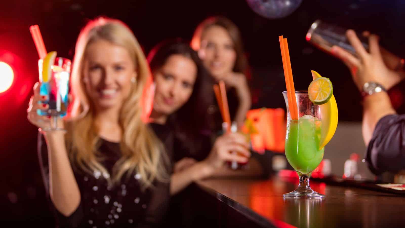 A group of ladies drinking cocktails with the focus of the image being the green cocktail in front.