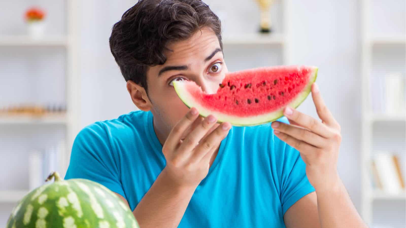 A man looking at the slice of watermelon he's holding with a bite taken out of it.