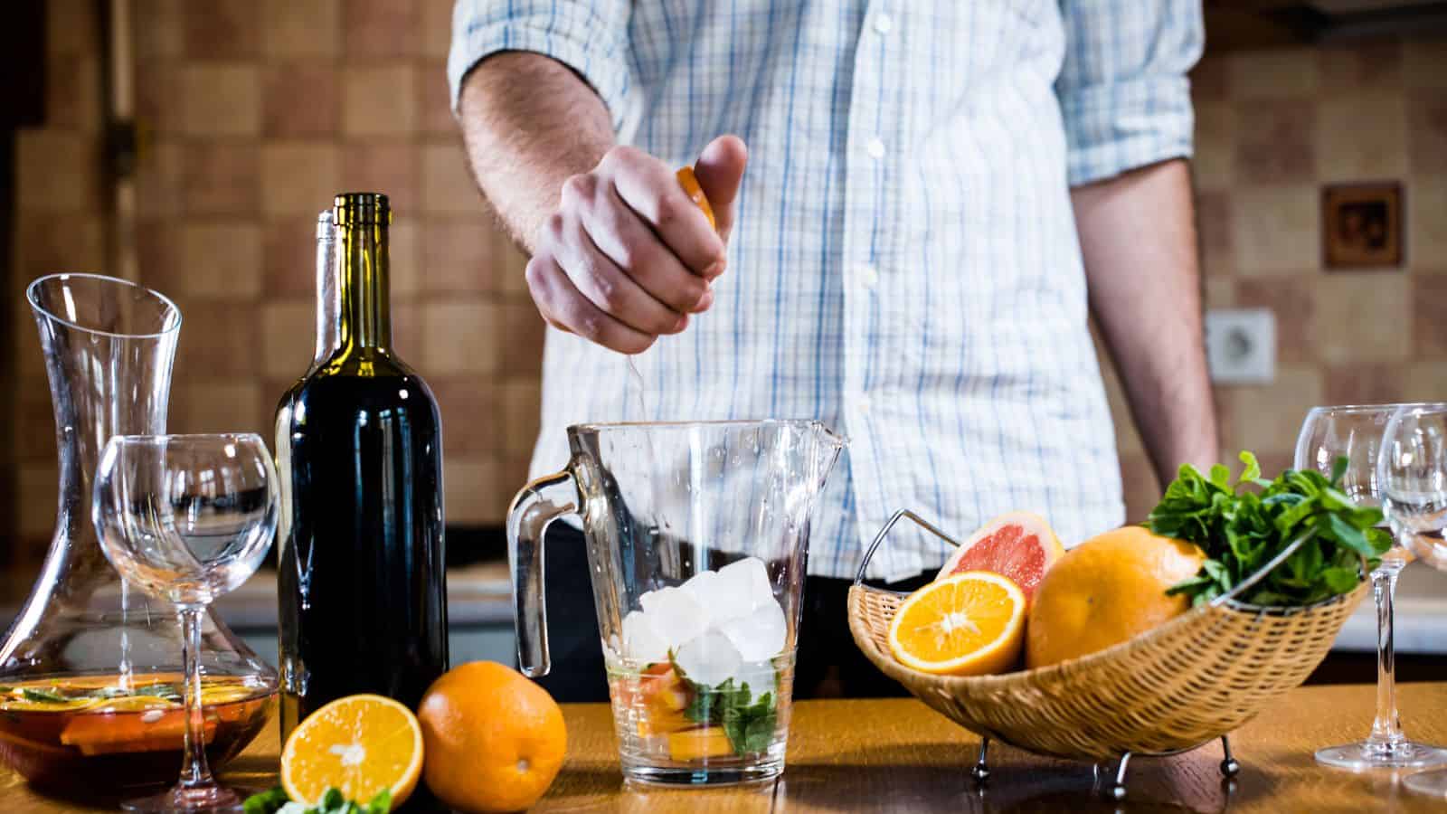 A person in a plaid shirt is squeezing an orange over a pitcher filled with ice and mint. A bottle of wine, two empty glasses, a bowl of arugula, and a basket of assorted citrus fruits are on the table.