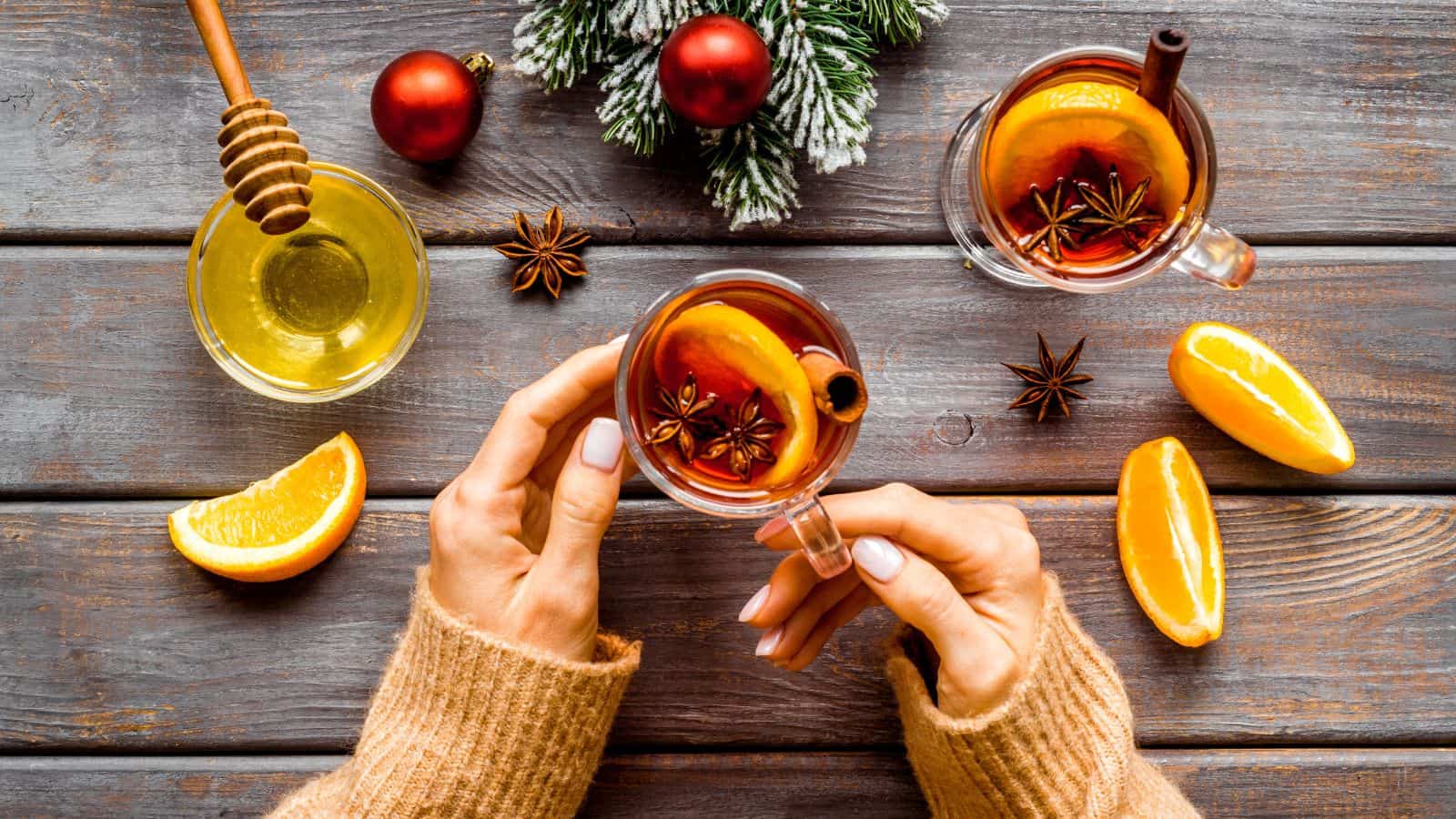 Two hands hold clear mugs of spiced tea with orange slices and cinnamon sticks. A wooden table features orange wedges, a honey dipper in honey, star anise, and holiday ornaments with pine branches.